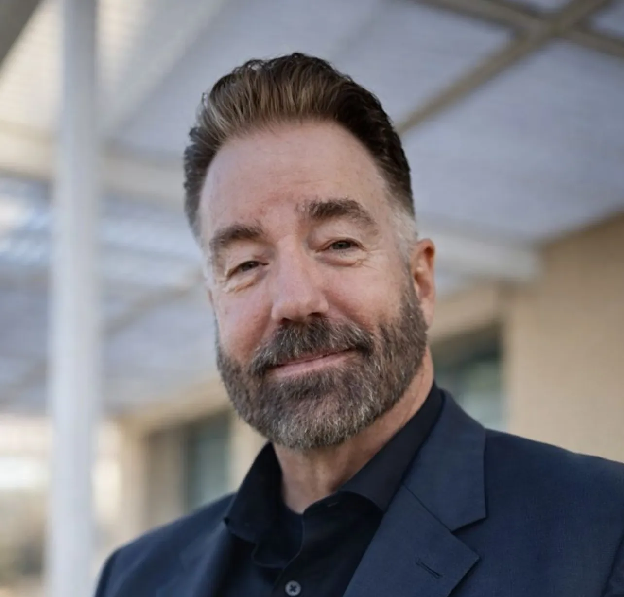 Close-up portrait of a middle-aged man with a beard and short hair, wearing a dark blue blazer and shirt, smiling slightly outdoors with a blurred building and white pillar in the background.