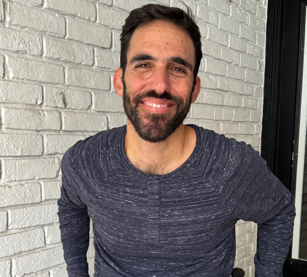 Smiling man with dark hair and beard wearing a dark blue long-sleeve shirt standing in front of a white brick wall.
