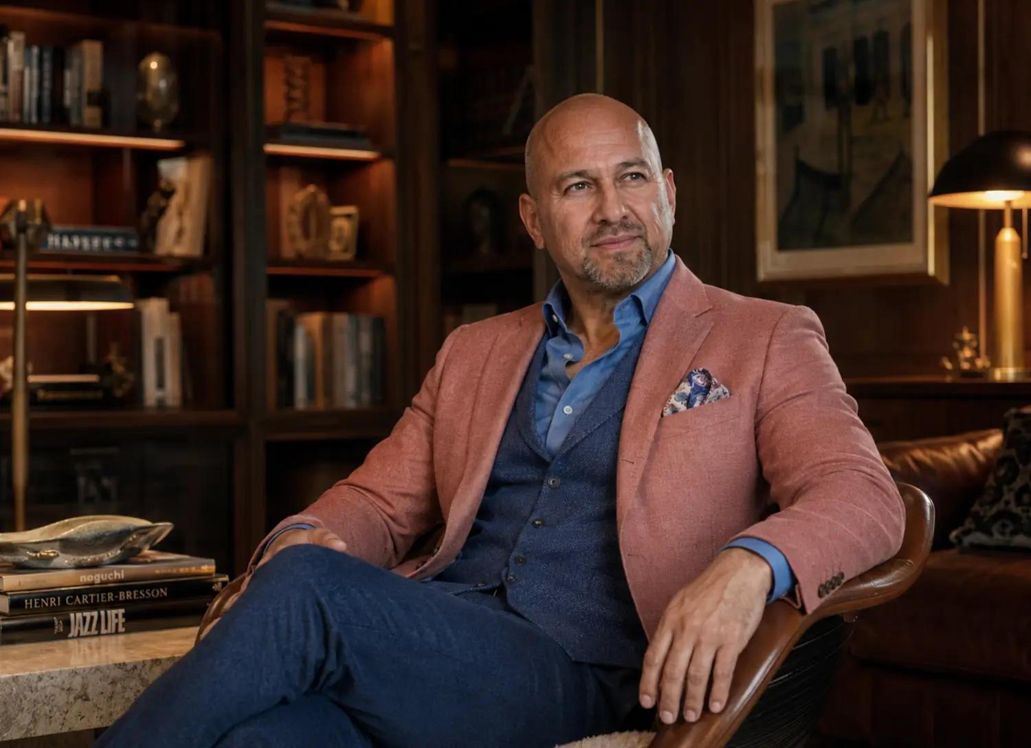 Bald middle-aged man with a trimmed beard wearing a dark double-breasted blazer with gold buttons and a white shirt, standing on a boat with the ocean and sky in the background.