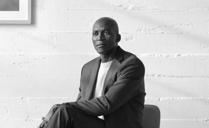 Black-and-white photo of a man with a shaved head wearing a blazer and white shirt, sitting with his hands resting on his knee against a textured white wall.