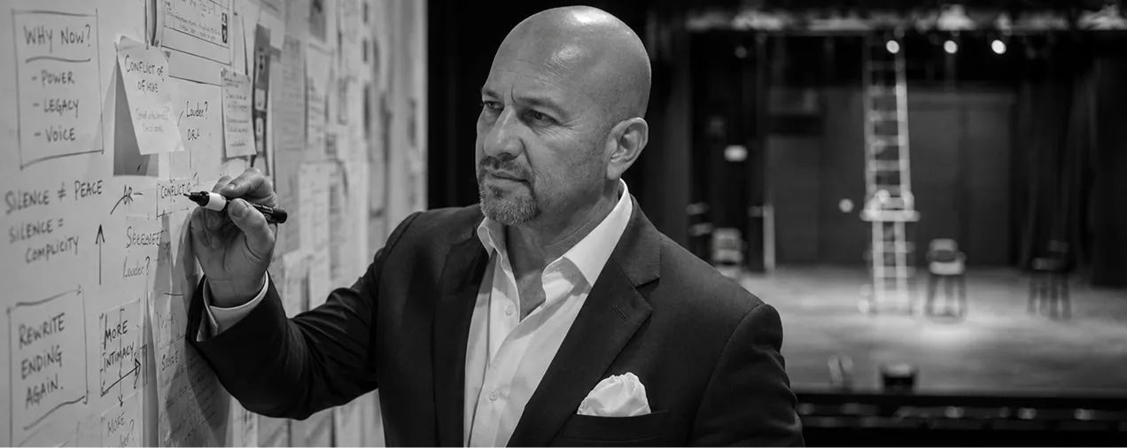 Man in a suit writing on a board filled with notes and diagrams, in a theater setting with empty chairs and a ladder in the background.