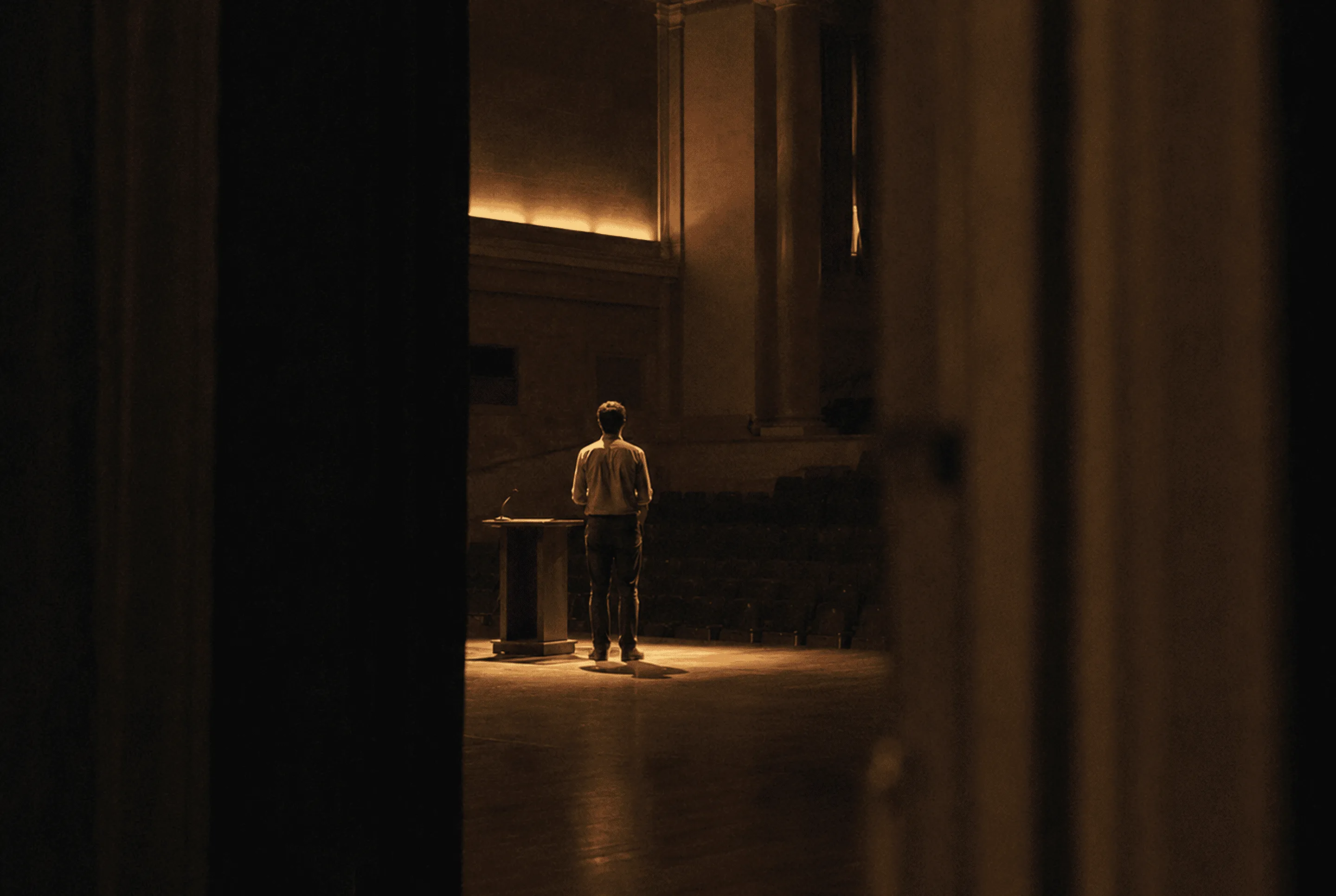 A person standing alone on a dimly lit stage behind a lectern with a microphone, viewed from a shadowed backstage area, facing an empty auditorium.