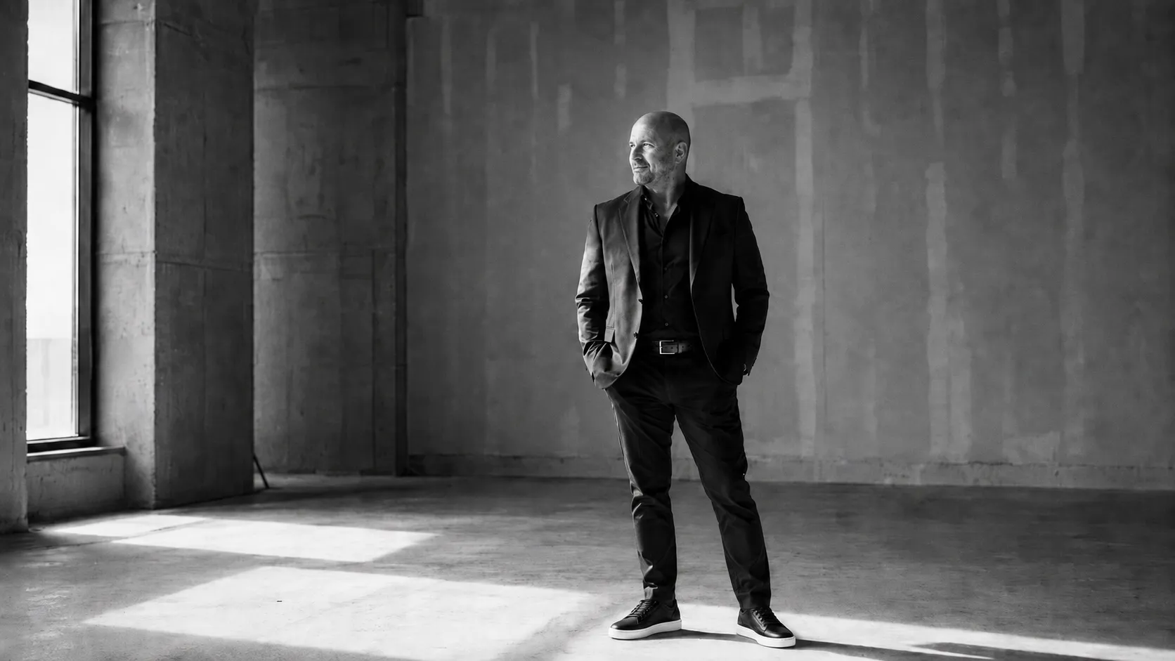 Black-and-white photo of a bald man wearing a dark suit and sneakers standing with hands in pockets in a large empty room with concrete walls and natural light from a window casting shadows on the floor.