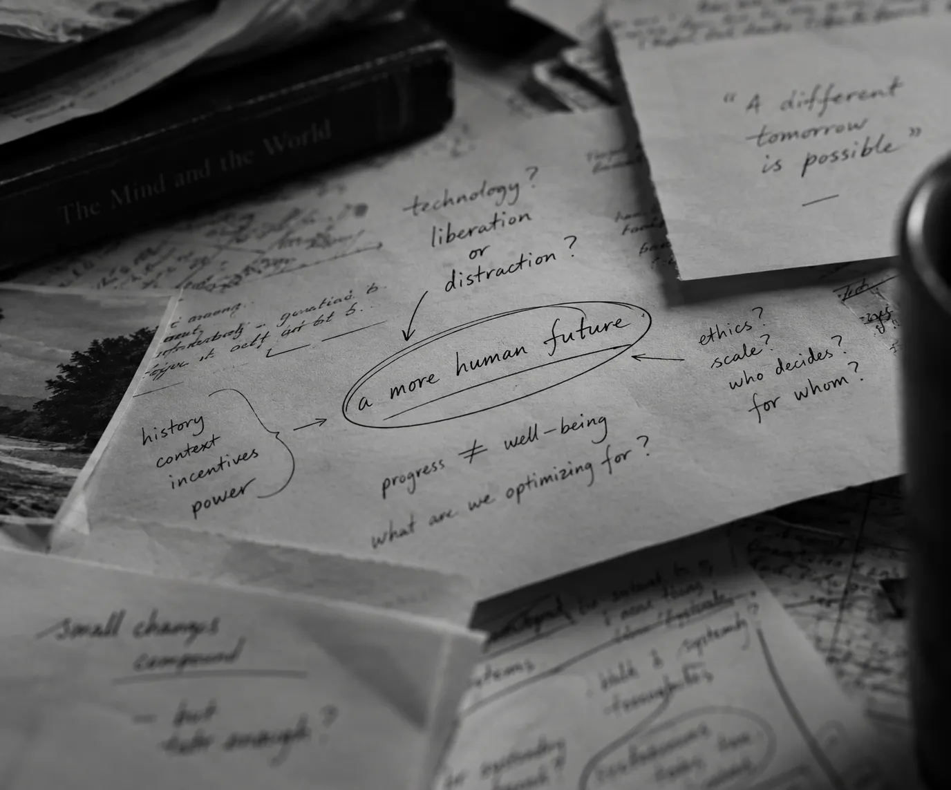 Black and white photo of handwritten notes on paper scattered on a desk with a book titled 'The Mind and the World.' Central note is circled and reads 'a more human future' with surrounding questions and phrases about technology, ethics, progress, and well-being.