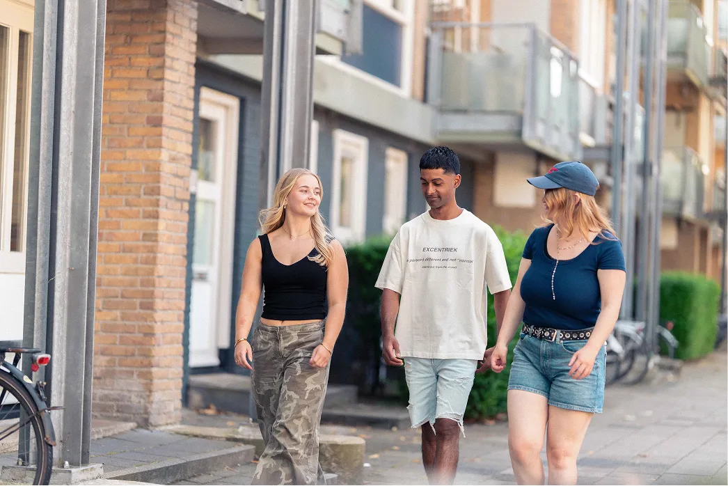 Drie jonge mensen wandelen buiten in casual kleding over een trottoir in een stad.
