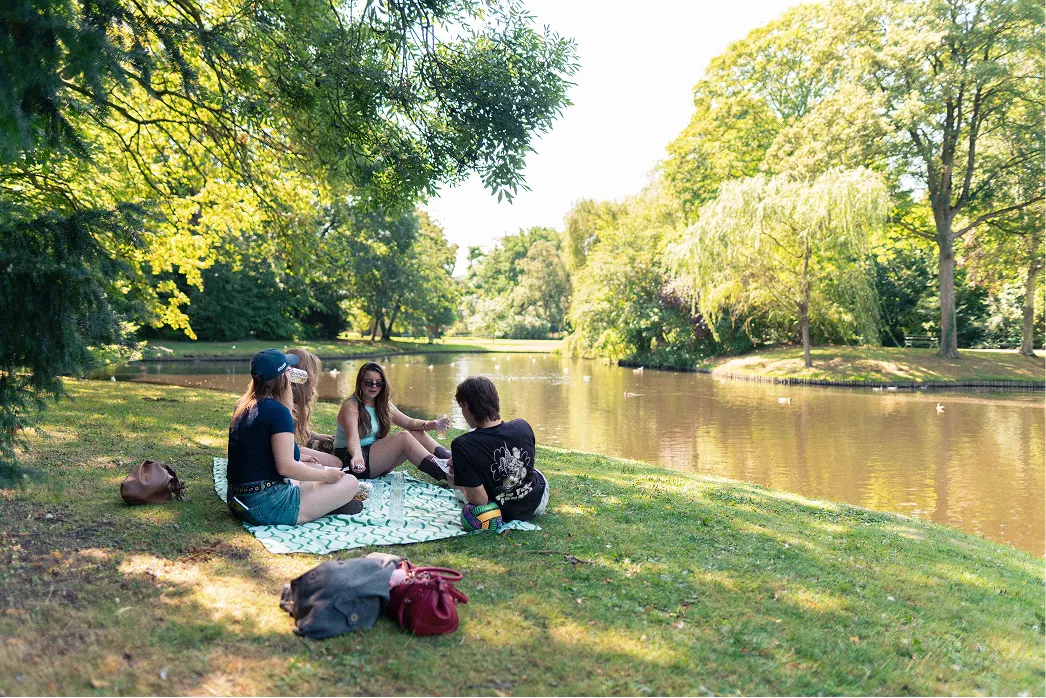 Een groep jongeren zit op een picknickkleed aan het water in het park en praat met elkaar.