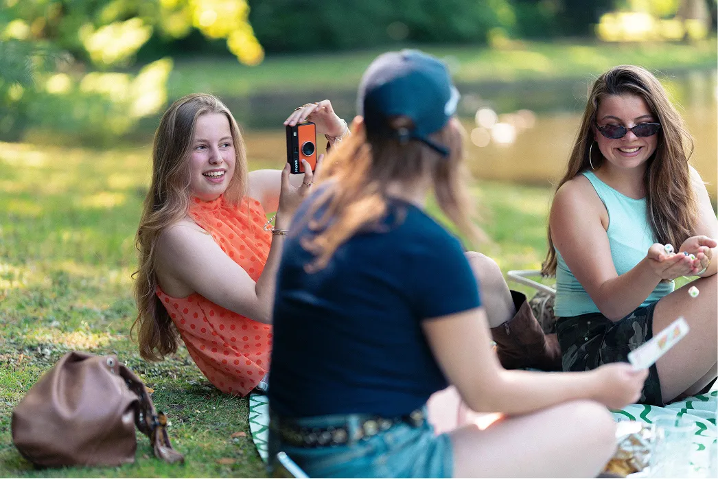 Een jongere maakt met een fotocamera een foto van twee vriendinnen in het park.