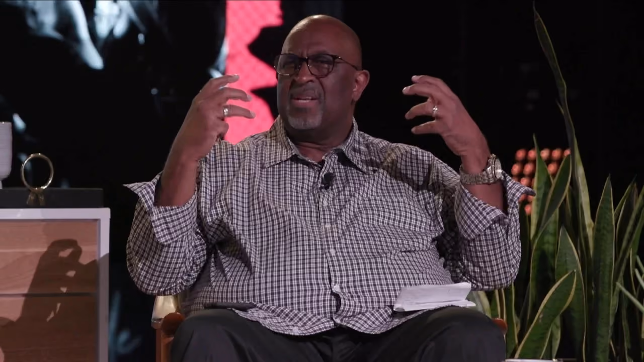 Man in checkered shirt gesturing while seated on talk show set.