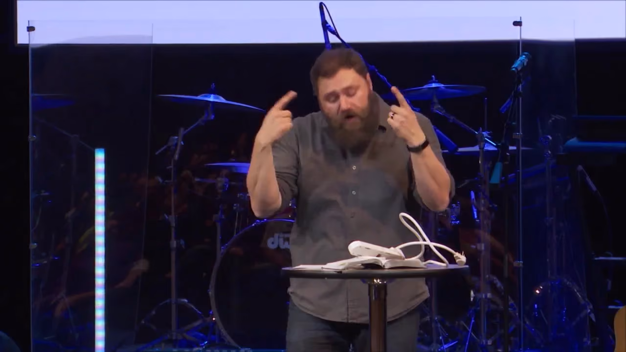 Man speaking at pulpit with drums behind him. He gestures with both hands. A book and wires rest on the table.