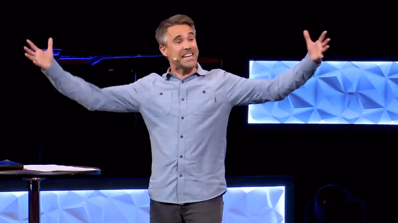 Man with arms wide open on a stage with blue geometric background.
