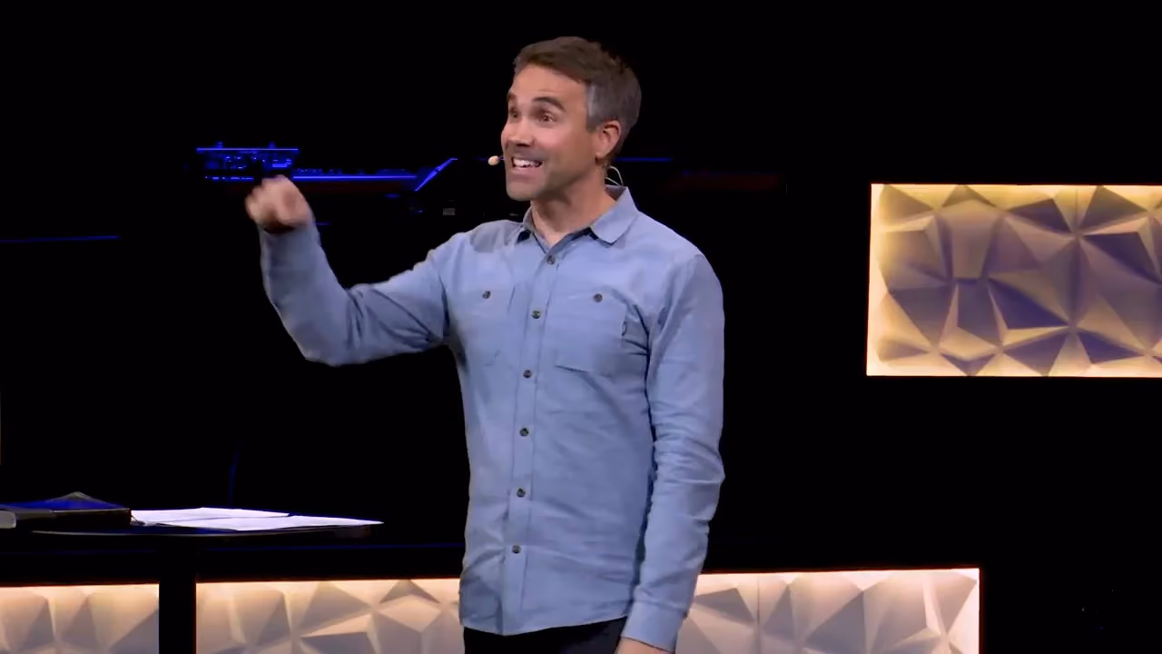 Man in blue shirt gesturing on stage with microphone visible. Lighted geometric panel behind him.