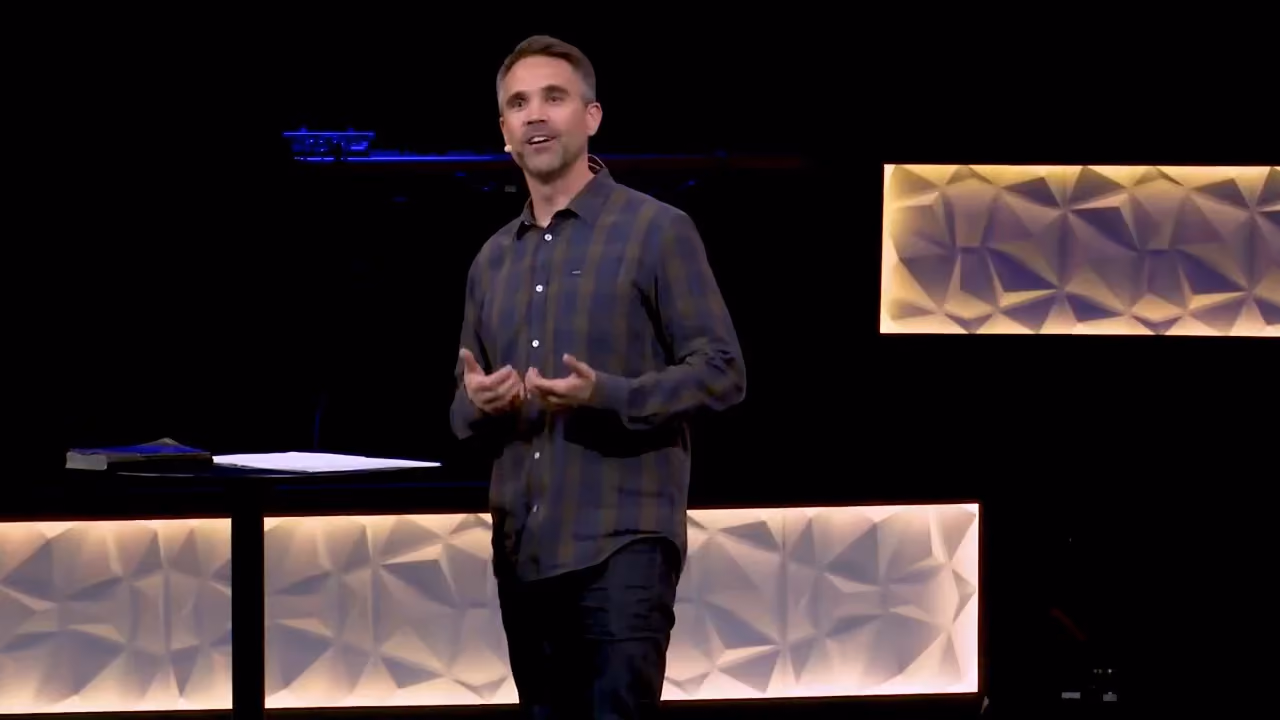 Man speaking on a dark stage with illuminated geometric panels behind him.