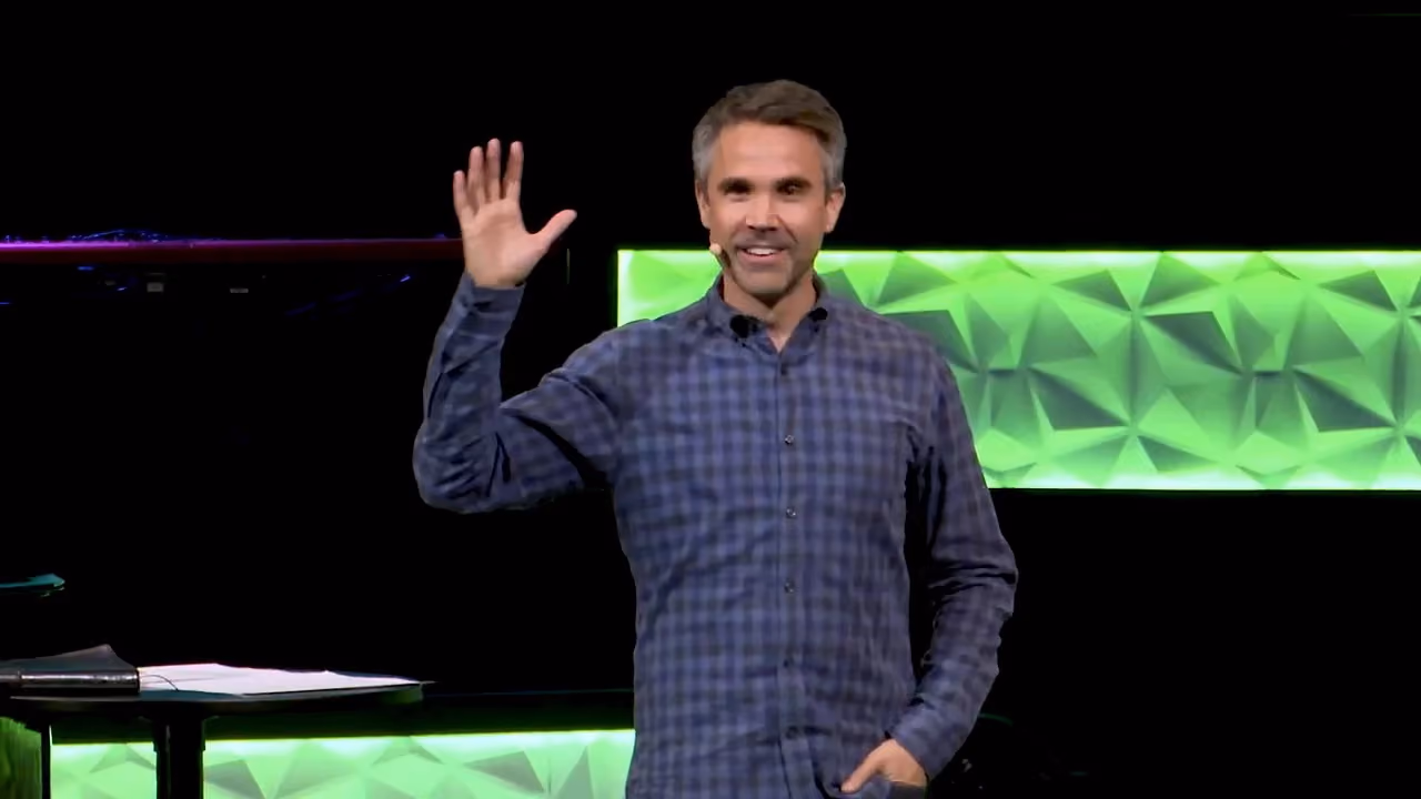 Man waving hand on stage with green geometric background. He wears a plaid shirt and has a microphone.
