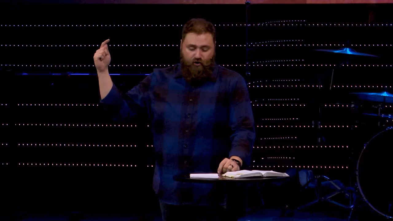 Man with beard speaking at podium with open book and pointing hand. Blue lighting and drum set visible in background.