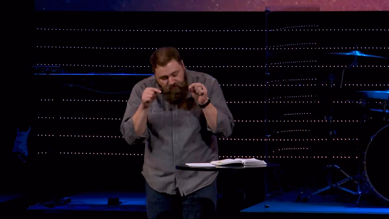 Man with beard speaking on stage with book on table behind him.