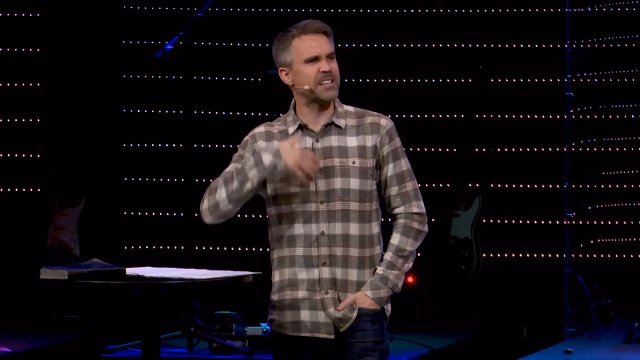 Man in plaid shirt speaking on stage with hand near chest and other hand in pocket.