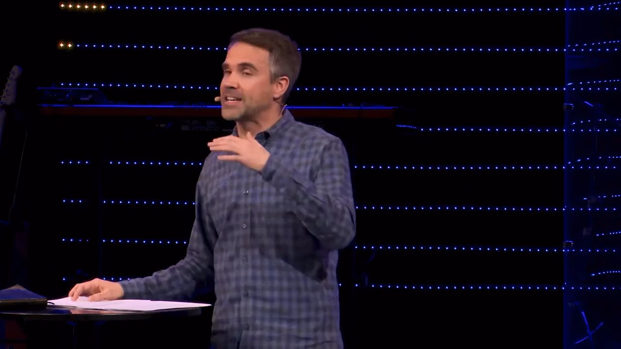 Man speaking on stage with hand gesture near chest holding papers on podium.