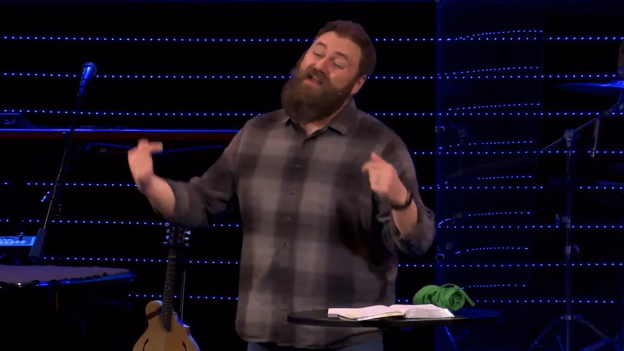 Man with beard speaking on stage with blue LED lights behind him.