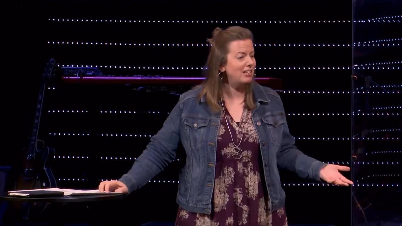 Woman in denim jacket speaking on stage with illuminated background.