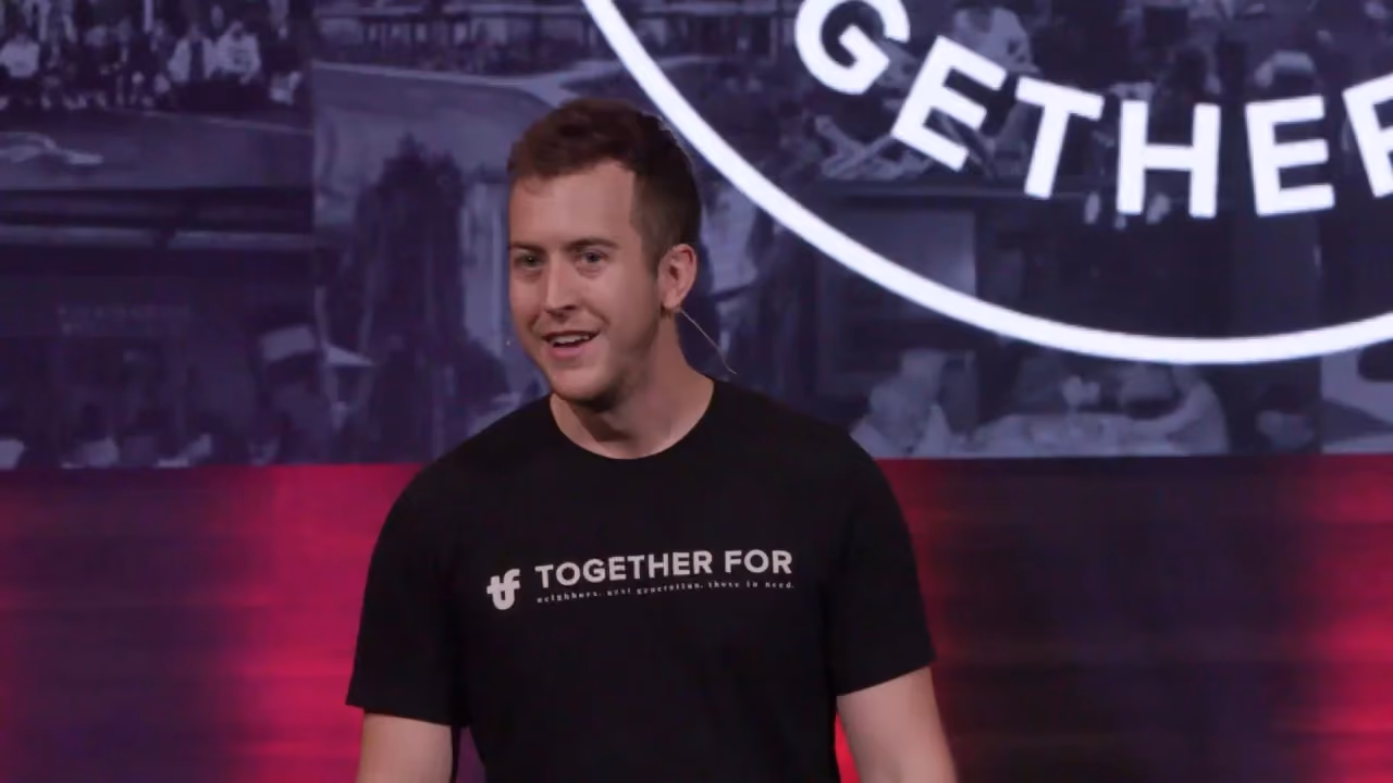 Man in black t-shirt speaking at event with 'Together For' logo on shirt and backdrop.