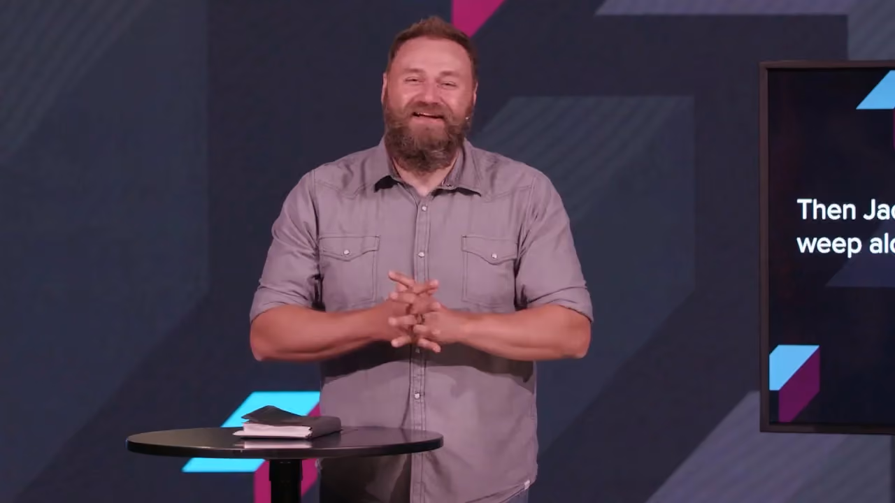 Man with beard speaking at podium with screen behind him.