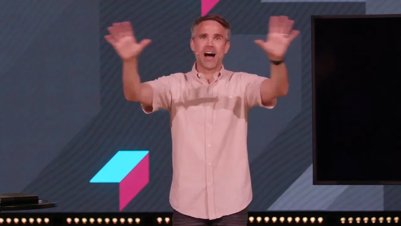 Man speaking with arms raised on a stage with geometric background design.