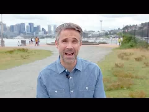 Man speaking outdoors near waterfront with city skyline in background.