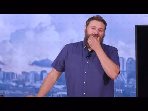 Man with beard touching chin in blue shirt against cityscape background.