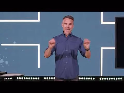 Man speaking on stage with raised hands in front of a blue background with white lines.