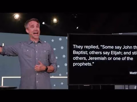 Man speaking on stage with Bible verse displayed on screen.