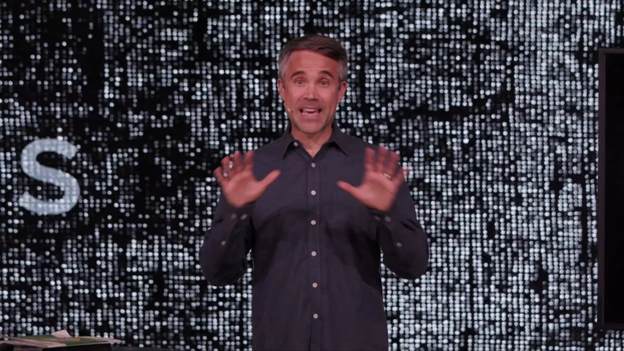 Man speaking with hands open in front of a pixelated screen with a large letter S visible.