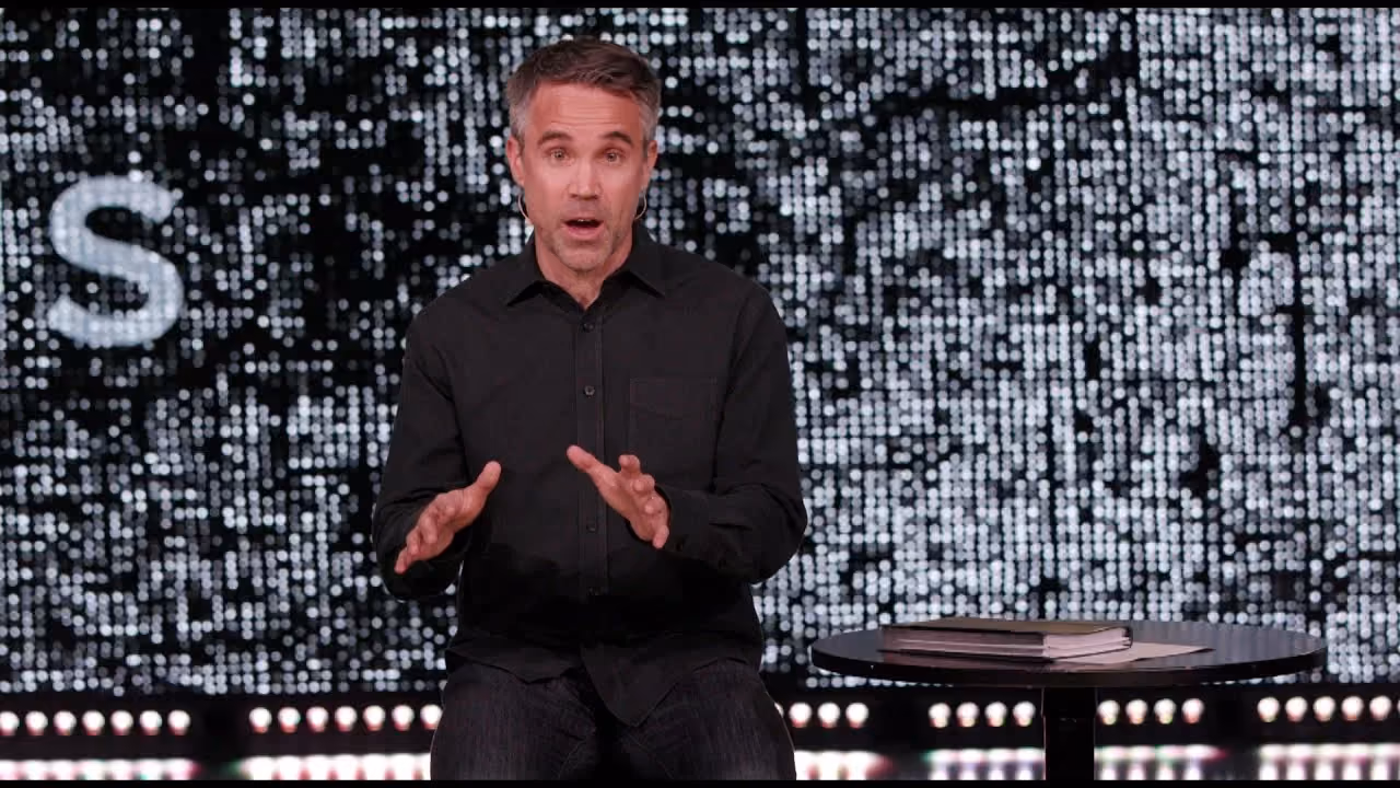 Man speaking on stage with digital pixelated backdrop and table with book.