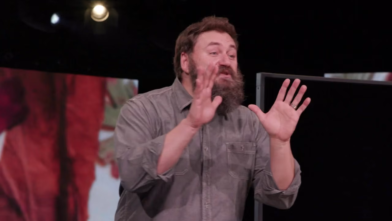 Man with beard gesturing with both hands raised. He wears a button-up shirt. Background includes a screen and red graphic.
