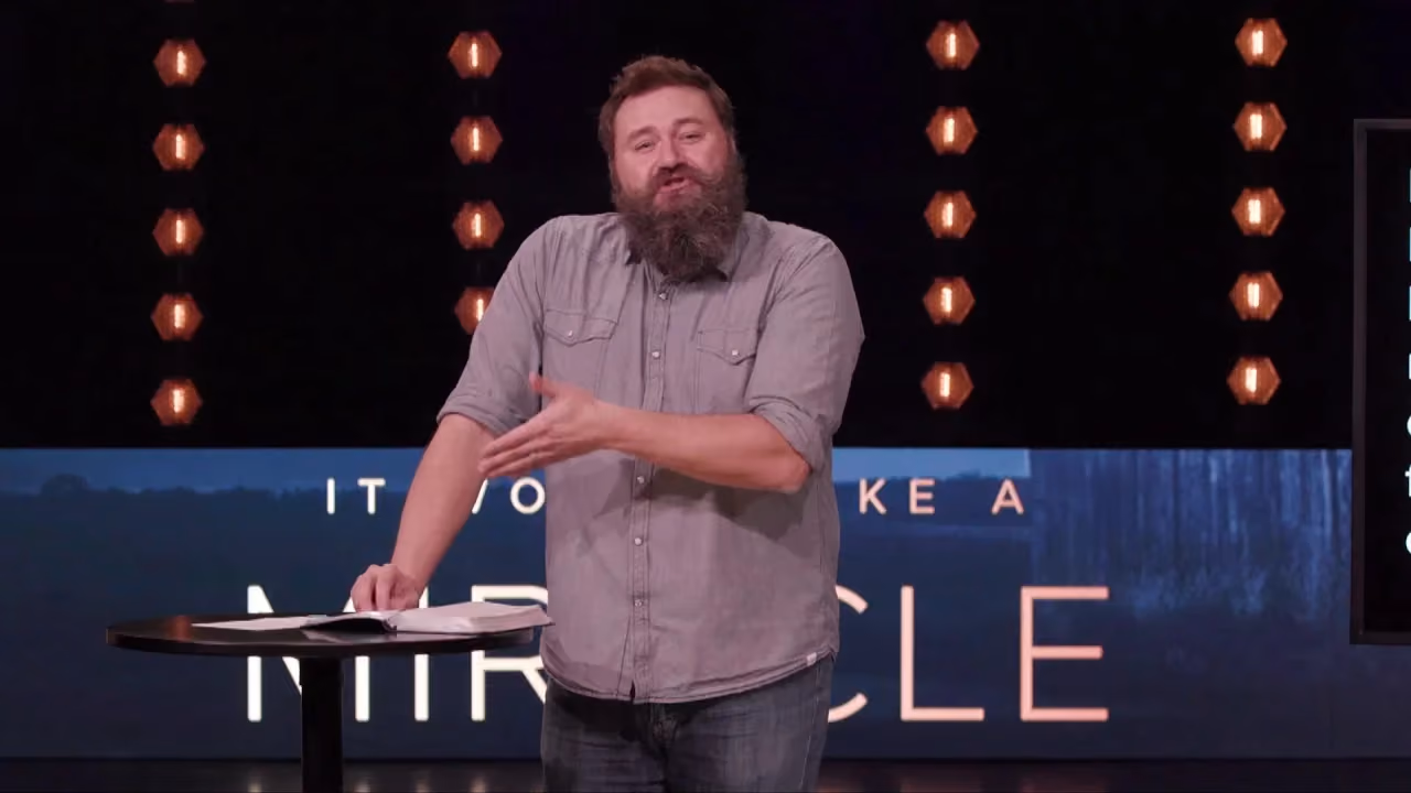 Man with beard speaking on stage with book on table behind him. Large letters read 'IT TO MAKE A MIRACLE'.