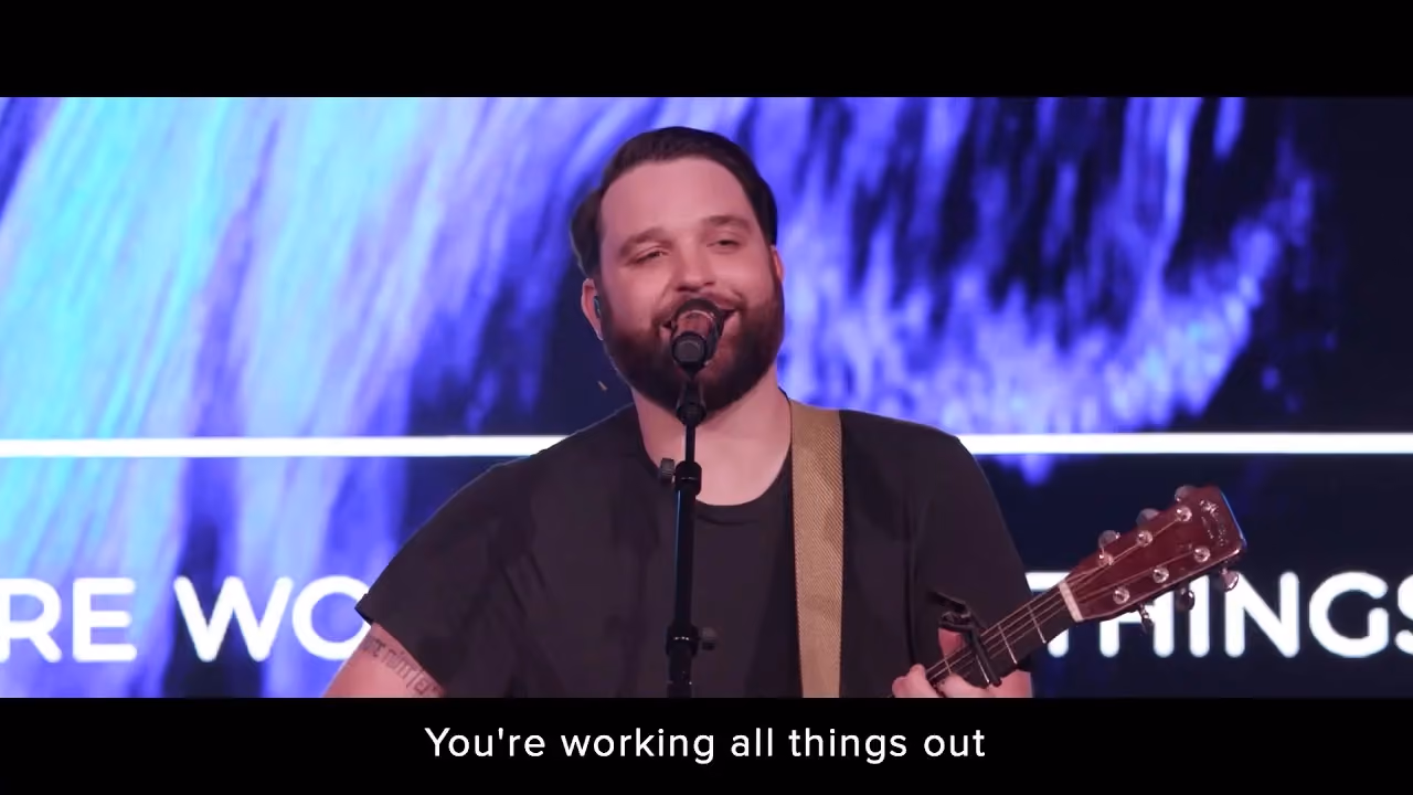 Man singing into microphone holding guitar on stage with blue background lighting.