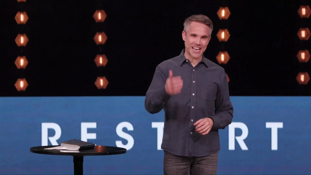 Man giving thumbs up on stage with 'RESTART' backdrop and table with book.