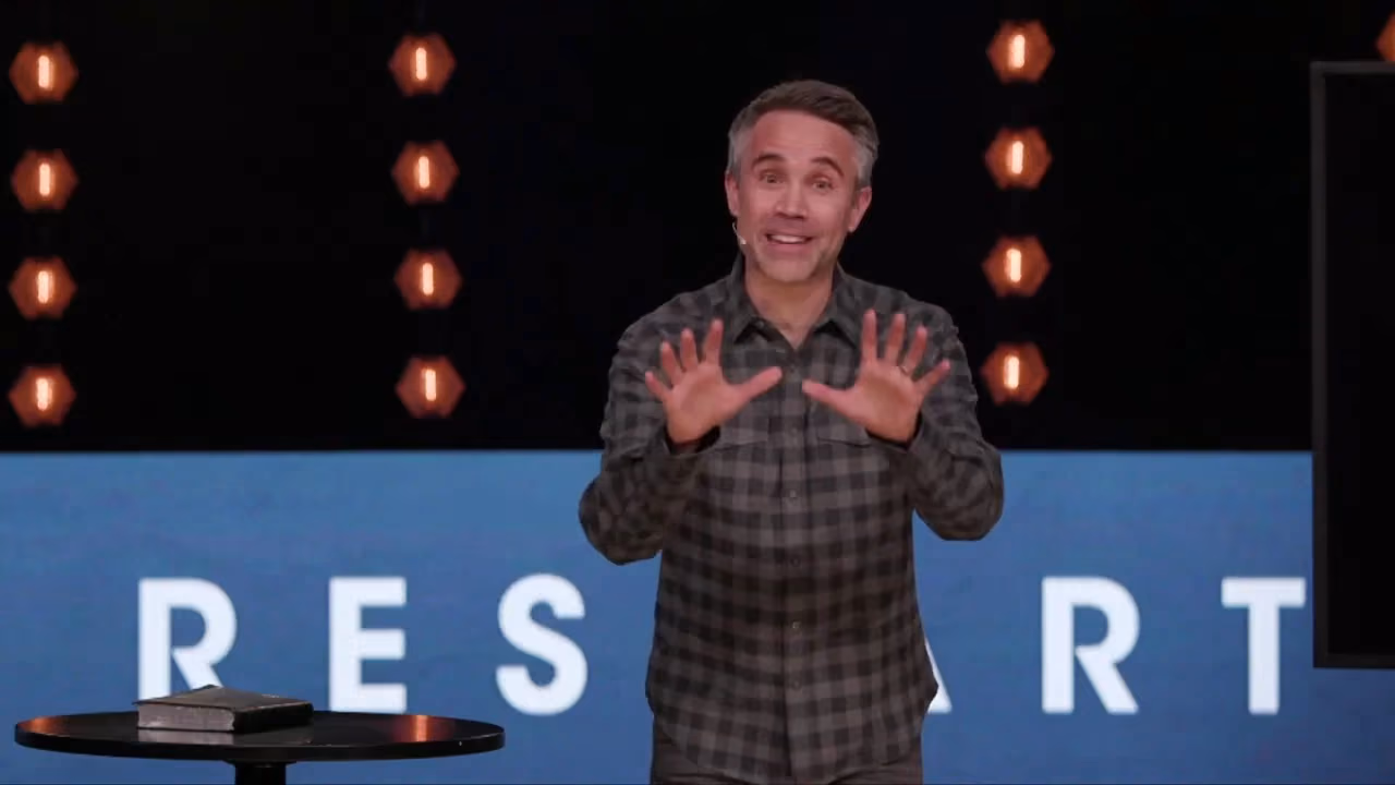 Man gesturing with hands open on stage in front of blue background with letters R E S A R T.