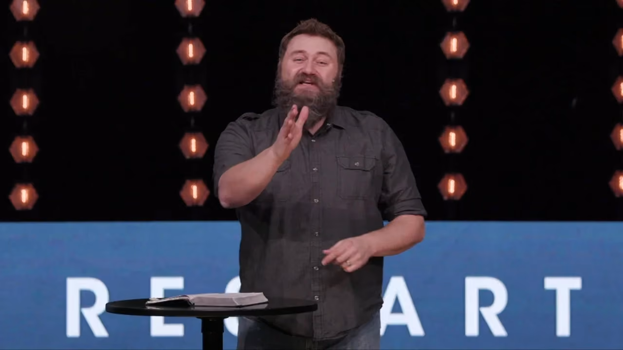Man with beard speaking at podium with book and illuminated background text.