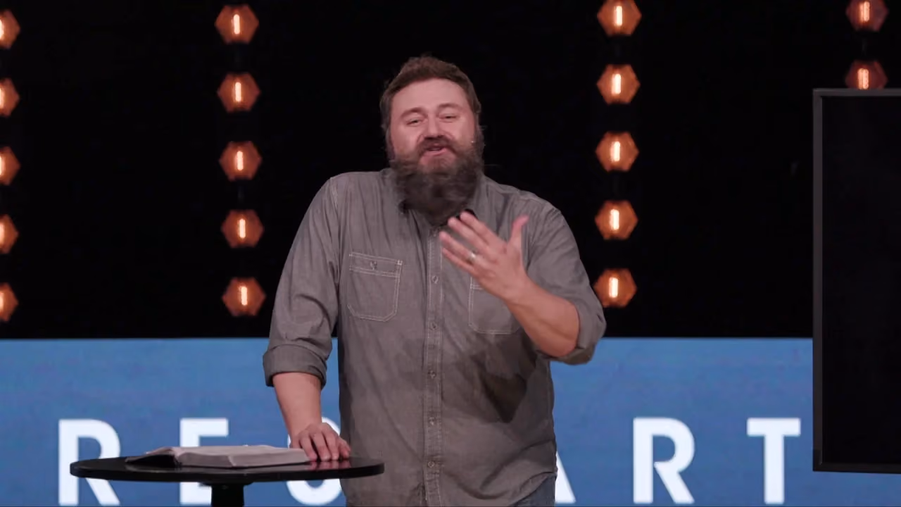 Man with beard speaking at podium with book and lights behind him.