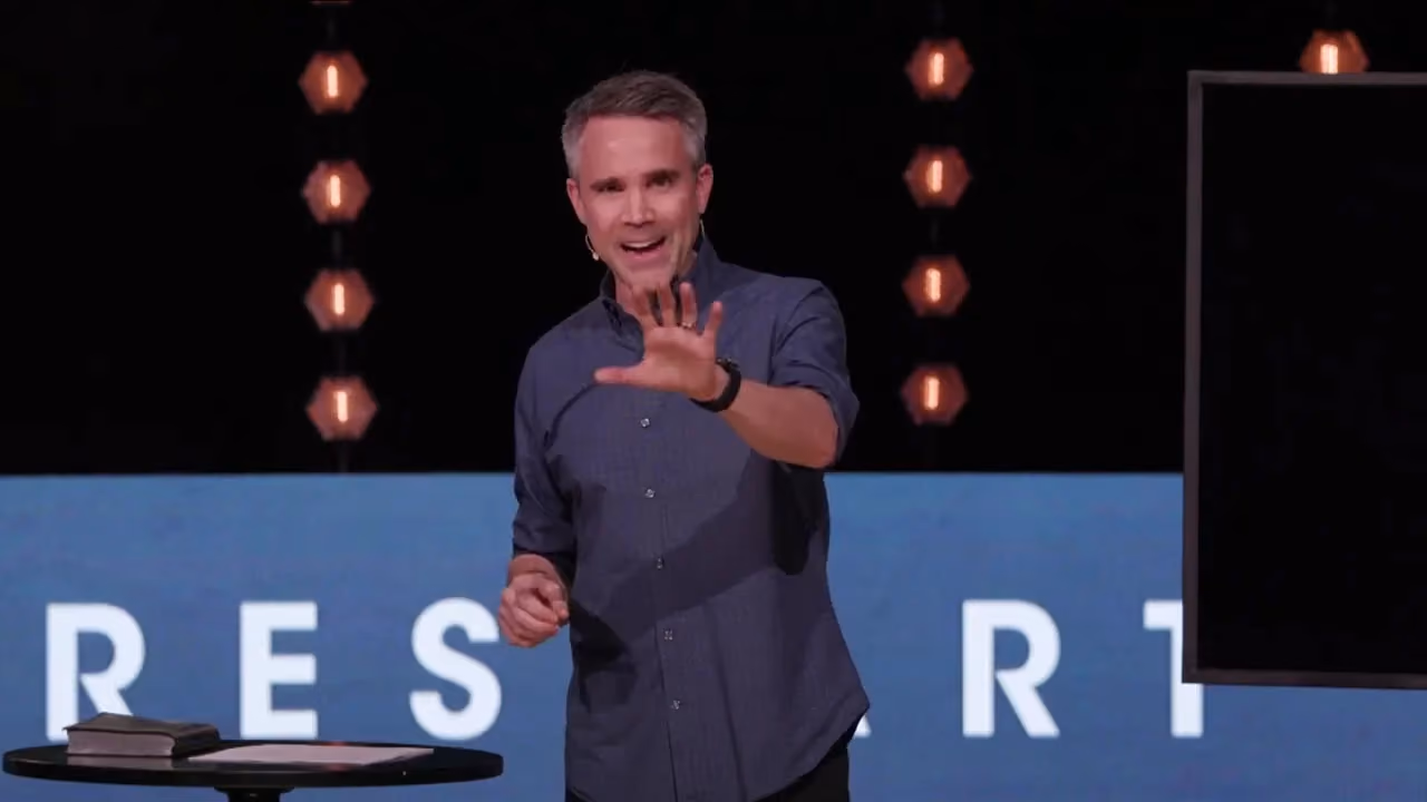 Man gesturing on stage with large letters behind him.