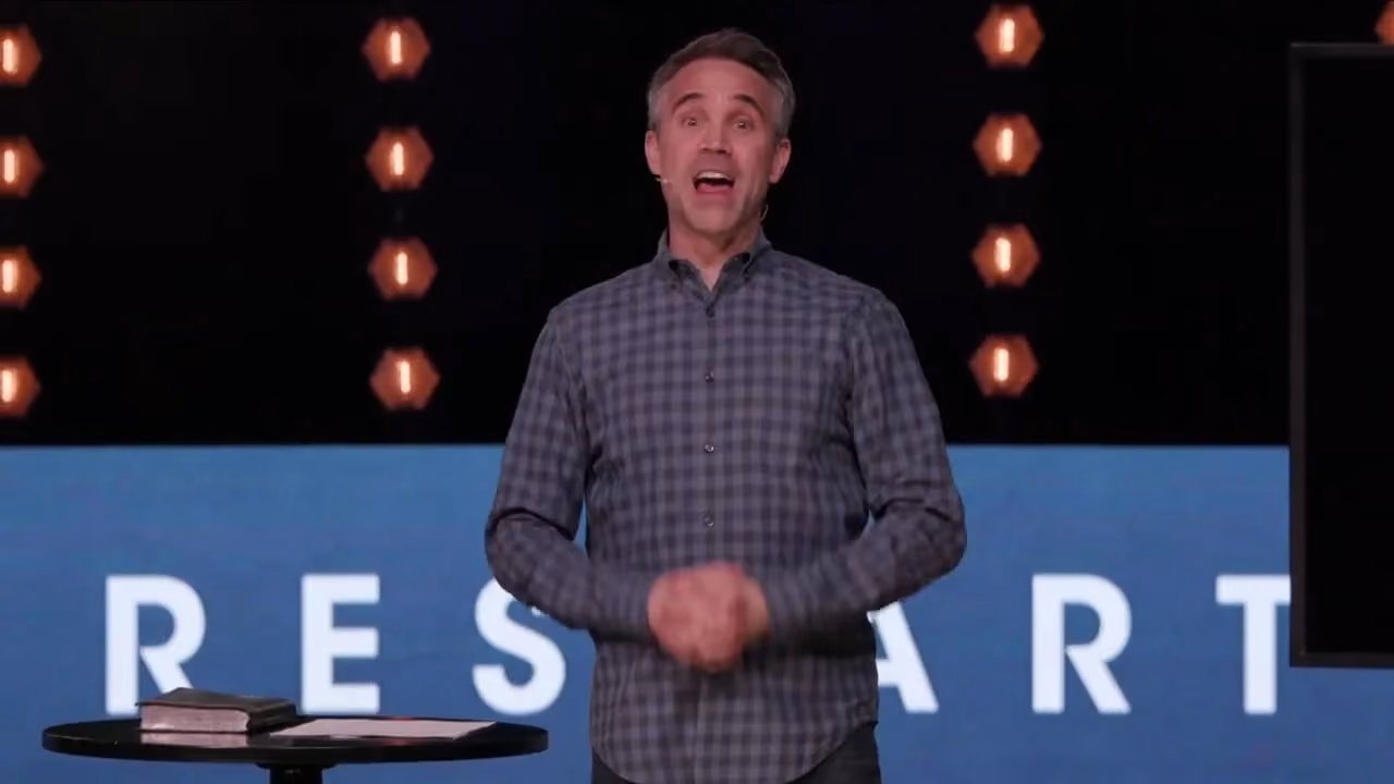 Man speaking on stage with 'RESTART' backdrop and hexagonal lights.