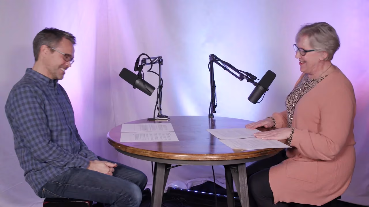 Two people sitting at a table with microphones and papers. One is smiling. Background is purple.