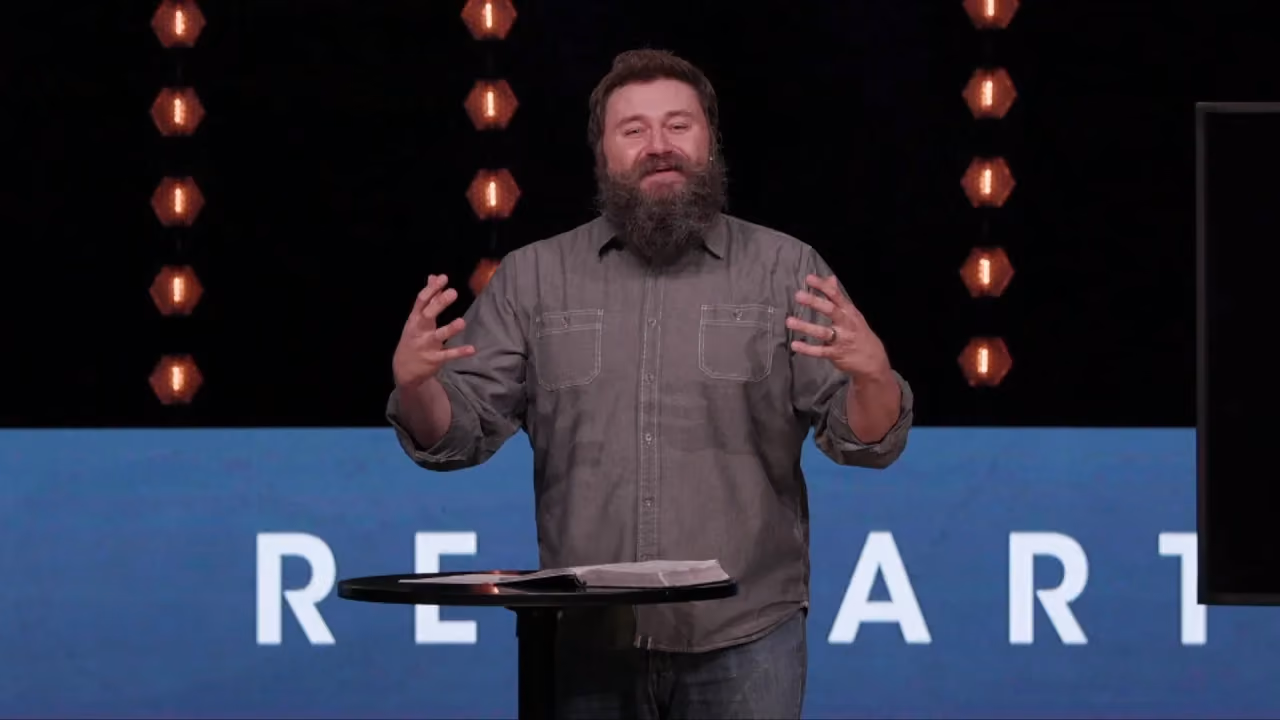 Man with beard speaking at podium with open hands. Large letters behind him. Lights above.