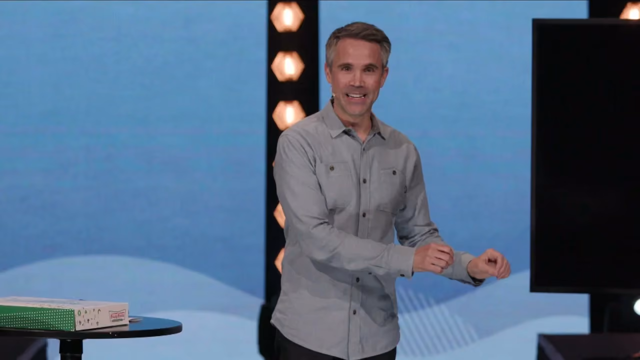 Man in gray shirt smiling on stage with blue background and lights.
