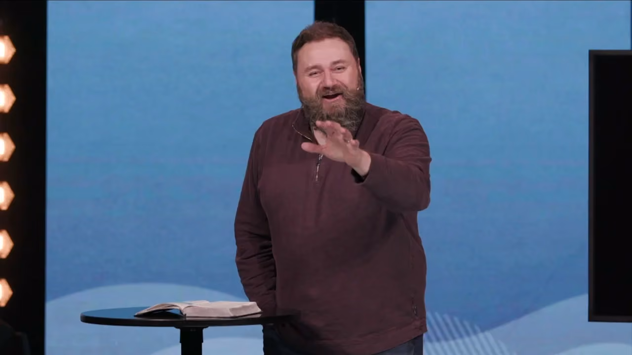 Man with beard gesturing while standing beside a small table with an open book.