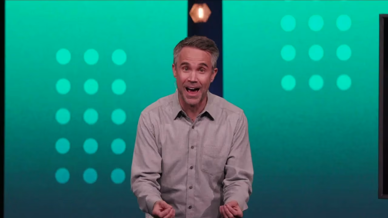 Man speaking on stage with green background and circular lights.