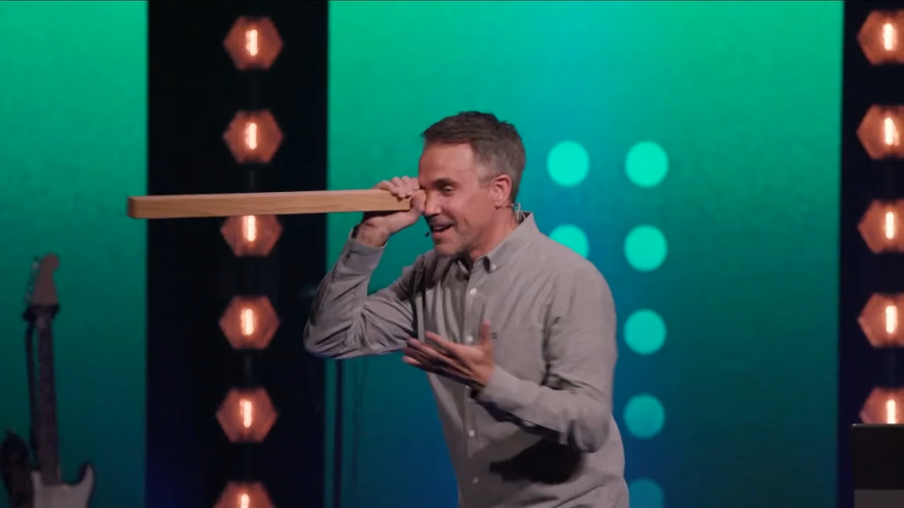 Man holding wooden bar to eye on stage with green background and lights.