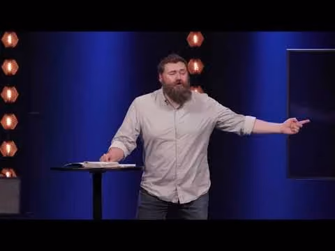 Man with beard speaking at podium with book and lights behind him.