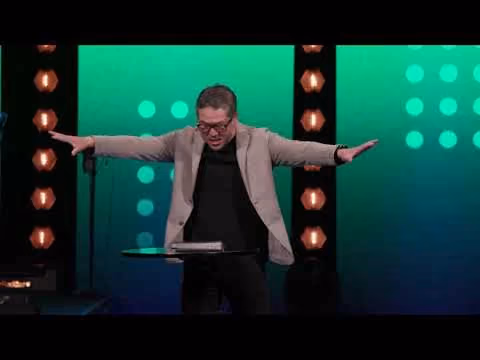 Man in beige jacket and glasses stands on stage with arms outstretched in front of green backdrop with lights.