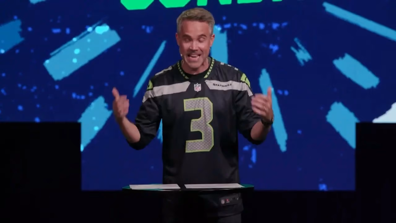 Man in black jersey with number three gesturing while speaking at podium. Blue background with abstract shapes.